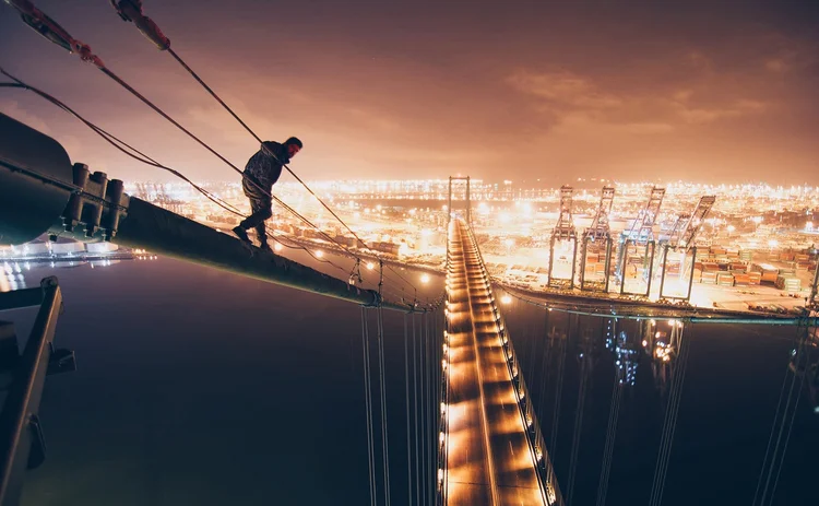 man-standing-on-steel-cable-of-suspension-bridge-over-river-by-harbor-at-night