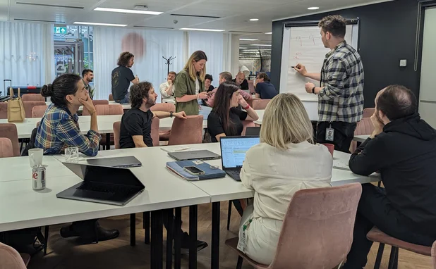 A team sat around a table facing someone writing on a whiteboard.