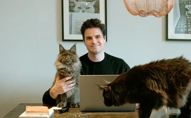 Andrew Leal sitting at his desk with one of his cats in his arm and the other standing on the desk.