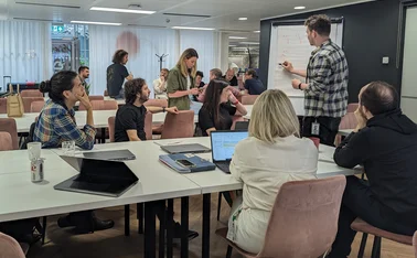 A team sat around a table facing someone writing on a whiteboard.