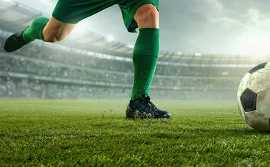 Footballer about to kick ball in floodlit stadium, while opponent attempts sliding tackle  