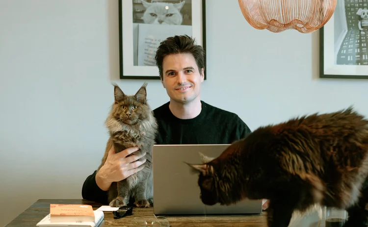 Andrew Leal sitting at his desk with one of his cats in his arm and the other standing on the desk.
