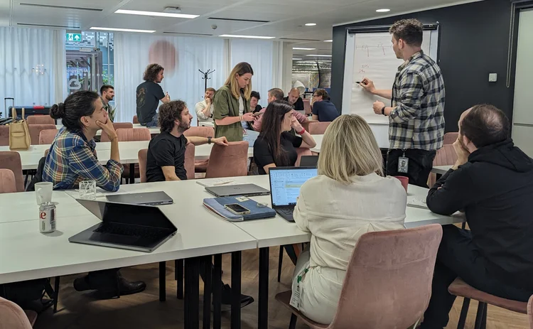 A team sat around a table facing someone writing on a whiteboard.