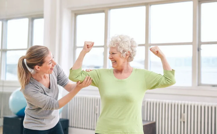 A lady administering rehabilitation to an older lady who has her arms raised in a victory pose