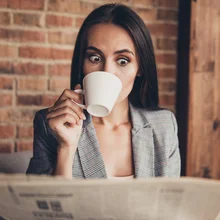 Close up portrait of astonishment brunette woman with crazy emotion and stare open eyed read the news about high oil prices drink espresso beverage from a white small mug in loft workstation