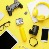 Overhead view of consumer electronics and gaming accessories including a gamepad, headphones, tablet, and action camera on a bright yellow background