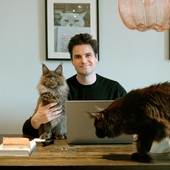Andrew Leal sitting at his desk with one of his cats in his arm and the other standing on the desk.