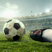 Footballer about to kick ball in floodlit stadium, while opponent attempts sliding tackle  