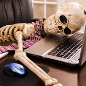 Skeleton of a man with head on laptop in his office. This dedicated employee has worked himself to death!
